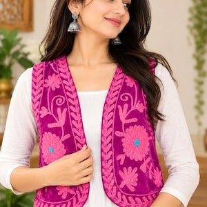 Op de afbeelding: Een fel magenta mouwloos vest met ingewikkeld roze bloemenborduurwerk en een klein felblauw accent in het midden van elke bloem. Het vest wordt gedragen over een effen wit shirt met lange mouwen en heeft een gedetailleerde bladrand langs de voorzijde.