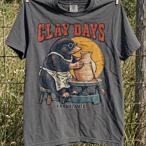 May include: A charcoal gray short-sleeve t-shirt featuring a graphic of a mole wearing glasses and an apron, sitting on a stool while shaping a clay pot on a pottery wheel. The text CLAY DAYS appears in orange above, and HAND FORMED is below.
