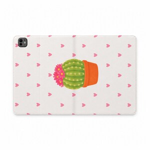 Può includere: Una custodia per tablet bianca con un motivo ripetuto di piccoli cuori rosa. Al centro della custodia è presente l'illustrazione di un cactus verde rotondo in un vaso arancione, decorato con un grappolo di fiori rosa acceso sul lato sinistro.