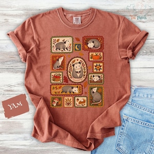May include: A terracotta-coloured short-sleeve t-shirt featuring a graphic print of multiple rectangular frames containing illustrations of opossums, pumpkins, mushrooms, and autumn leaves. The central oval frame highlights a single opossum.