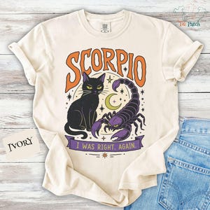 以下が含まれることがあります： アイボリーのTシャツで、オレンジ色の文字でSCORPIOと書かれ、その下に黒い猫と紫色のサソリが描かれたグラフィックプリントが特徴です。黄色い三日月と星が動物たちを囲んでいます。下の紫色のバナーにはI WAS RIGHT. AGAIN.と記されています。