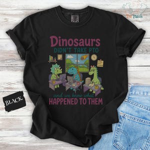 Op de afbeelding: Een zwart t-shirt met een afbeelding van drie dinosaurussen die aan bureaus werken. De tekst luidt: Dinosaurs DIDN'T TAKE PTO and we know what HAPPENED TO THEM. De illustratie bevat computers, een raam met een meteoor en kantoorbenodigdheden.