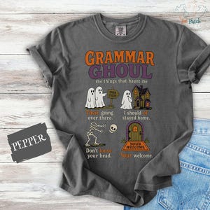 May include: A charcoal gray t-shirt featuring the text Grammar Ghoul and the things that haunt me. The graphic includes ghosts, a haunted house, a skeleton, and a door with text corrections for common grammar mistakes like their, of, loose, and your.