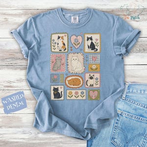 May include: A light blue t-shirt featuring a graphic print of various cats in rectangular and heart-shaped frames, interspersed with small flowers and gingham patterns. The shirt has a crew neckline and short sleeves, with the text WASHED DENIM.