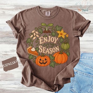 May include: An espresso brown t-shirt featuring a graphic print of a wreath made of green vines, a sprout, seeds, a yellow flower, a slice of pumpkin pie, and two pumpkins, one of which is a jack-o'-lantern, with the text "ENJOY every SEASON".