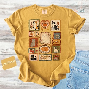 Puede incluir: Una camiseta de color amarillo mostaza con un estampado gráfico central de varios gatos, calabazas, hojas de otoño, setas y una taza de café dispuestos en una cuadrícula de marcos rectangulares y en forma de corazón.