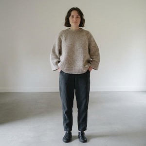 Peut inclure: Un pull en tricot beige à grosses mailles avec un col rond côtelé et des manches larges, associé à un pantalon gris anthracite foncé avec des coutures verticales et des baskets basses noires. Le pull porte une petite étiquette blanche rectangulaire.