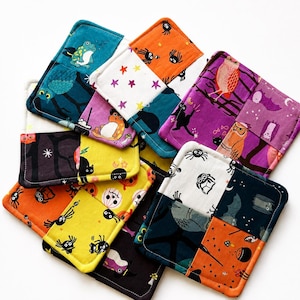 May include: A collection of square fabric coasters featuring patchwork designs with Halloween-themed patterns including owls, bats, spiders, pumpkins, cats, and stars in vibrant colors like orange, purple, yellow, teal, black, and white.