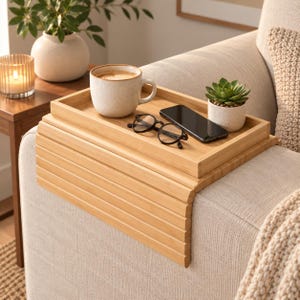 Peut inclure: Un plateau d'accoudoir de canapé flexible en bambou avec une surface plane rectangulaire et des côtés à lattes qui se drapent sur l'accoudoir. Le plateau contient une tasse en céramique avec du café, un smartphone noir, des lunettes et une succulente.