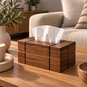 Puede incluir: Una caja rectangular para pañuelos hecha de madera de color marrón oscuro con un patrón de veta visible. La caja presenta un diseño segmentado en bloques con ranuras verticales y horizontales. Un pañuelo de papel blanco sobresale.