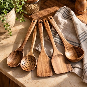 Peut inclure: Un ensemble de cinq ustensiles de cuisine en bois présentant un motif de grain de bois brun naturel. La collection comprend deux cuillères pleines, une cuillère à trous avec des perforations circulaires et deux spatules plates.