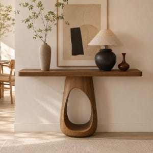 Peut inclure: Une console en bois dotée d'un plateau rectangulaire lisse et d'un pied central unique, creux et en forme de goutte. La table est décorée d'un grand vase beige contenant des branches vertes, d'une lampe noire et d'un petit vase marron.