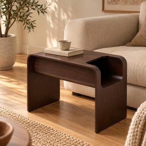 Peut inclure: Une table d'appoint en bois brun foncé avec un cadre incurvé en forme de C et un compartiment de rangement ouvert intégré sur le côté droit. Un petit bol en céramique de couleur claire repose sur un livre rectangulaire sur la table.