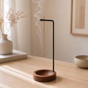 Op de afbeelding: Een verticale wierookhouder met een ronde voet van donker hout en een dunne, zwarte metalen arm die omhoog steekt en buigt om een brandend wierookstokje vast te houden, met een dunne sliert witte rook die van de punt afkomt.
