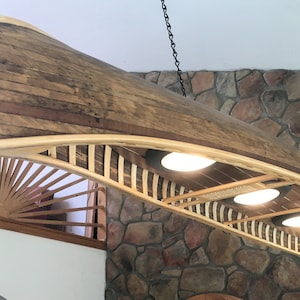 May include: A wooden canoe-shaped light fixture with a rustic finish hangs from a chain. The canoe is illuminated by four light bulbs. The fixture is suspended from a stone wall.