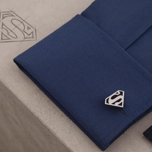 Puede incluir: Un gemelo de metal plateado con un emblema en forma de diamante y una letra S estilizada, sujeto al puño doblado de una camisa azul oscuro. Un boceto a juego del emblema con la letra S es visible en la superficie cercana.