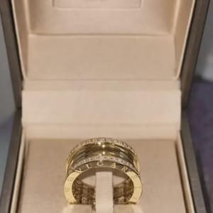 Könnte beinhalten: Ein goldener Bvlgari-Ring mit einem mehrreihigen Design und Pavé-Diamanten an den oberen und unteren Rändern. Das mittlere Band ist mit dem Markennamen Bvlgari graviert. Der Ring befindet sich in einer grauen Schmuckschatulle mit beigem Futter.