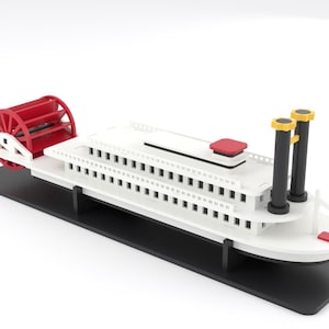Puede incluir: Un modelo de barco de vapor blanco con una rueda de paletas trasera roja, dos chimeneas negras con puntas amarillas y un acento rojo en la cubierta. La embarcación está montada sobre una base rectangular negra, con una estructura de niveles.