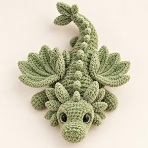 May include: A sage green crocheted dragon plush toy featuring textured wings, a long tail with a fin, and a row of small spikes running down its back. The creature has large, dark, glossy eyes and a detailed head with multiple soft, rounded protrusions.
