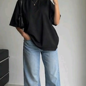 May include: A black short-sleeved t-shirt with a crew neckline paired with light blue wide-leg denim jeans. The outfit includes a small black shoulder bag, a gold chain necklace, and white sneakers, creating a casual ensemble.