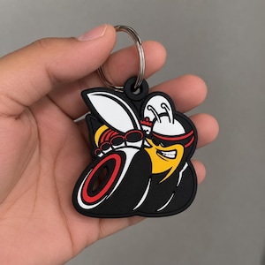Può includere: Un portachiavi in gomma raffigurante un personaggio a forma di vespa stilizzata nei colori giallo, nero e bianco, con una fascia rossa sulla testa e un'espressione determinata. Il design presenta un'ala bianca prominente, un corpo a strisce gialle e nere e un dettaglio circolare rosso.