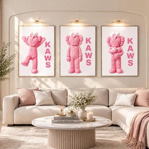 May include: Three rectangular wall art prints featuring pink fuzzy KAWS character figures on a white background. Each print displays the text KAWS in vertical pink capital letters. The figures are depicted in different poses, including standing and waving.