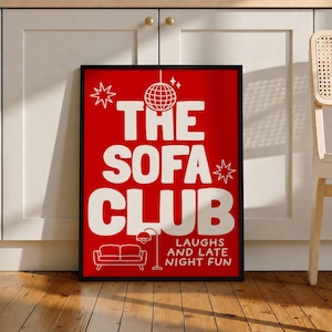 May include: A rectangular framed wall art print with a vibrant red background and bold white text that reads THE SOFA CLUB. The design includes a white disco ball, star icons, a floor lamp, a sofa illustration, and the text LAUGHS AND LATE NIGHT FUN.