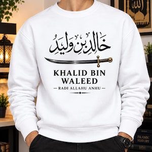 May include: A white long-sleeve crewneck sweatshirt featuring black Arabic calligraphy at the top, a graphic illustration of a curved sword in the center, and the text KHALID BIN WALEED followed by RADI ALLAHU ANHU with decorative lines below.