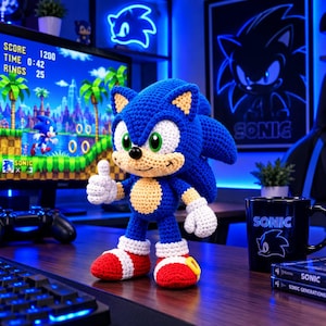 Op de afbeelding: Een handgemaakte gehaakte Sonic the Hedgehog knuffel met blauw garen, een beige buik, witte handschoenen en rode sneakers met gele gespen. Het personage staat met een duim omhoog op een bureau voor een computermonitor.