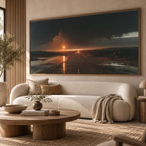 May include: A large rectangular wall art print featuring a landscape of a long, wet road stretching towards a bright sunset under dark, dramatic storm clouds. The scene is framed in a thin wooden border and displayed above a cream-coloured sofa.