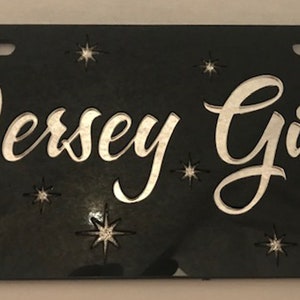 May include: Black license plate with silver lettering that reads "Jersey Girl". The plate is decorated with silver glitter stars.