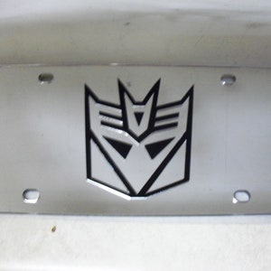 May include: A silver license plate frame with a black and silver Transformers Autobot symbol.