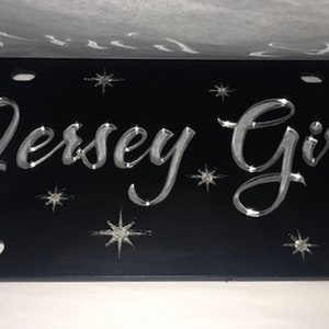 May include: Black license plate with silver lettering that reads "Jersey Girl" and silver star accents.