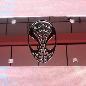 May include: A silver license plate with a black Spider-Man mask design. The mask is cut out of the metal and has a web pattern.