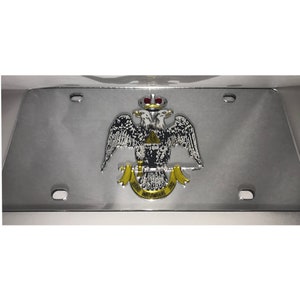 May include: A silver license plate frame with a gold and black emblem featuring two eagles, a crown, and a triangle with the letter 'G' inside. The text on the emblem reads 'Deus Meumque Jus'.