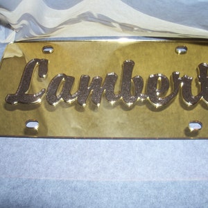 May include: Gold-colored license plate with the name "Lambert" in black cursive lettering.