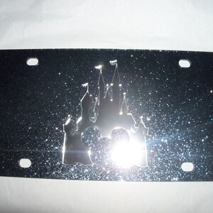 May include: A black license plate with a silver glitter finish. The plate features a cutout design of the iconic Disney castle with Mickey Mouse ears in the center.