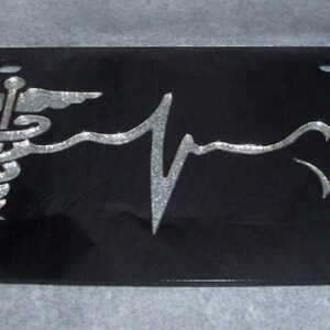 May include: Black license plate with a silver medical symbol, a silver heart, and a silver EKG line.