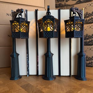 May include: Three black, 3D-printed street lamp bookends featuring intricate lattice patterns on the lantern housing. Each lamp contains a warm, glowing yellow LED light. The lamps are positioned in front of vertical books with white pages.