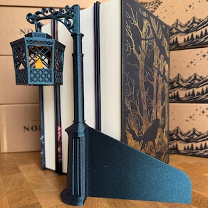 Könnte beinhalten: Ein schwarzer Buchständer in Form einer dekorativen Straßenlaterne mit einer gitterartigen Laterne, die ein kleines, gelb leuchtendes Licht enthält. Der Laternenpfahl steht auf einem flachen, dreieckigen Sockel zur Buchstütze mit strukturierter Oberfläche.