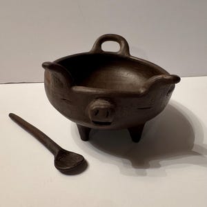 Peut inclure: Un bol en argile brun foncé sculpté à la main en forme de cochon, doté d'un museau, d'oreilles et d'une anse sur le bord. Le bol repose sur trois pieds courts et est accompagné d'une cuillère en argile brun foncé assortie à embout arrondi.