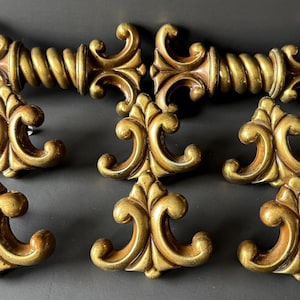 May include: A collection of eight gold-toned metal hardware pieces, including two elongated drawer pulls with a twisted rope centre and six smaller, symmetrical decorative knobs featuring curved, scroll-like flourishes and pointed tips.