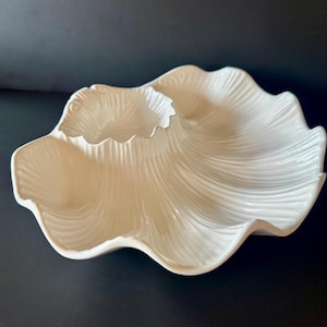 May include: A glossy off-white ceramic serving dish shaped like a large seashell. The piece features a central circular compartment surrounded by a larger, scalloped section, both decorated with fine, radiating ridges that mimic natural shell textures.