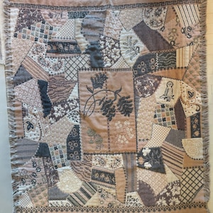 Peut inclure: Un plaid tissé rectangulaire présentant un motif patchwork dans des tons beige, marron et gris anthracite. Le design comprend diverses formes géométriques, des motifs floraux, des imprimés cachemire et une illustration centrale de pommes de pin.