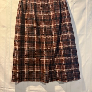 May include: A knee-length skirt featuring a brown, black, and cream plaid pattern. The skirt has a high-waisted design with a straight silhouette and a vertical slit at the bottom center front, hanging from a clear plastic garment hanger.