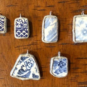 May include: Six pendants crafted from broken ceramic shards with blue and white patterns. Each piece is encased in a silver-toned metal frame with a small loop at the top for hanging. The shards feature various floral and geometric designs.