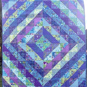 May include: A rectangular quilt featuring a geometric pattern of triangles and squares in shades of purple, blue, teal, and lime green. The fabric displays various floral, paisley, and abstract prints, all stitched together with a visible quilted texture.