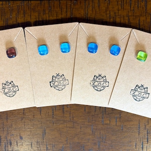 May include: Four pairs of square glass stud earrings displayed on individual brown cardboard cards. The earrings feature translucent colors including deep red, light blue, dark blue, and bright green. Each card is stamped with an L.B.C. logo.