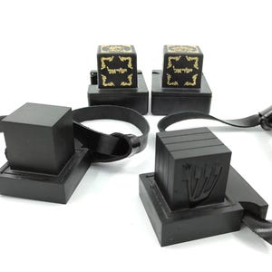 Puede incluir: Un conjunto de cajas de tefilín negras con correas de cuero. Dos cajas presentan texto hebreo dorado y bordes ornamentados, mientras que otra caja muestra la letra hebrea Shin en relieve en el lateral. Objetos de material negro mate.