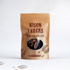 May include: A brown paper bag with a clear window showing a dark brown bison jerky product. The bag has the text "BISON TRACKS" in brown letters and the text "bison jerky with a kick" in smaller brown letters. The bag also has the text "BOURBON FLAVORED" in brown letters.